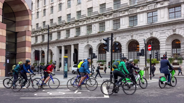 Several cyclists, dressed in work attire and helmets, ride across a city intersection in the Bank area of London